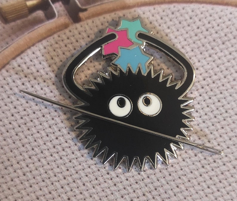 May include: A black enamel pin depicting a cartoon creature with a spiky body, large white eyes, and a needle through it. The pin has a silver border and holds three colourful star-shaped objects. The pin is set against a light purple fabric.