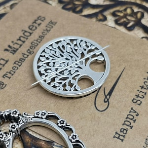 Könnte beinhalten: Ein silberner Fadenhalter mit einem Baum des Lebens-Design, durch dessen Mitte eine Nadel gefädelt ist. Der Fadenhalter liegt auf einer braunen Oberfläche mit dem Text "Mini Winders @TheBadgeshackUK" und "Happy Stitching" darauf.