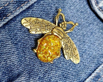 May include: A gold-colored bee-shaped brooch with intricate wing details and a shimmering, golden-yellow body. The brooch is set against a blue denim background.