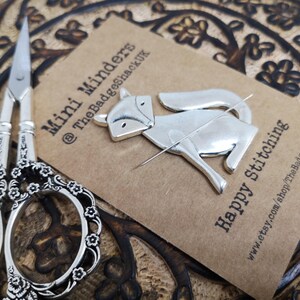 May include: Silver fox-shaped needle minder with a needle threaded through it. The needle minder is on a brown card with the text "Mini Minders @TheBadgeShackUK" and "Happy Stitching www.etsy.com/shop/TheBadgeShackUK".