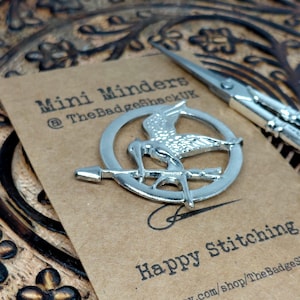 May include: A silver-colored metal pin featuring a bird design within a circle, resting on a brown card with the text "Mini Minders" and "Happy Stitching." A pair of silver scissors is also present. The background is a carved wooden surface.