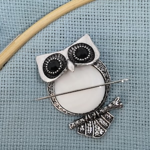 May include: A silver owl-shaped needle minder with black eyes and a white porcelain center. The needle minder is sitting on a blue Aida cloth.
