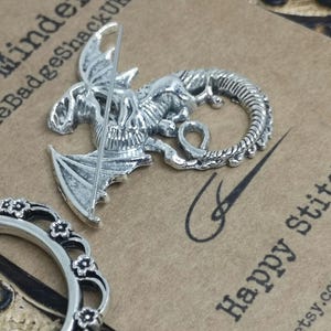 May include: Silver dragon needle minder with a circular ring. The dragon is curled around the ring, with its wings spread out. The needle minder is a decorative accessory for needlepoint, embroidery, and other crafts.