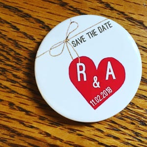 May include: White round save the date button with a red heart with the initials R & A and the date 11.02.2018. The heart is hanging from a piece of twine tied in a bow.
