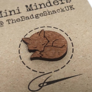 May include: A wooden fox pin in a curled up sleeping position. The pin is brown and has a white outline. The pin is on a brown background with the text "Mini Minders @TheBadgeShackUK".