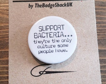 Może przedstawiać: Biały okrągły guzik z czarnym tekstem, który brzmi "Support Bacteria... they're the only culture some people have." Guzik jest przymocowany do brązowej kartki z tekstem "Needle Minder by TheBadgeShackUK" i adresem strony internetowej.