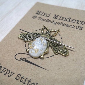 May include: A gold bee-shaped needle minder with a white and gold resin body. The needle minder is on a brown card with the text "Mini Minders @TheBadgeShackUK Happy Stitches".