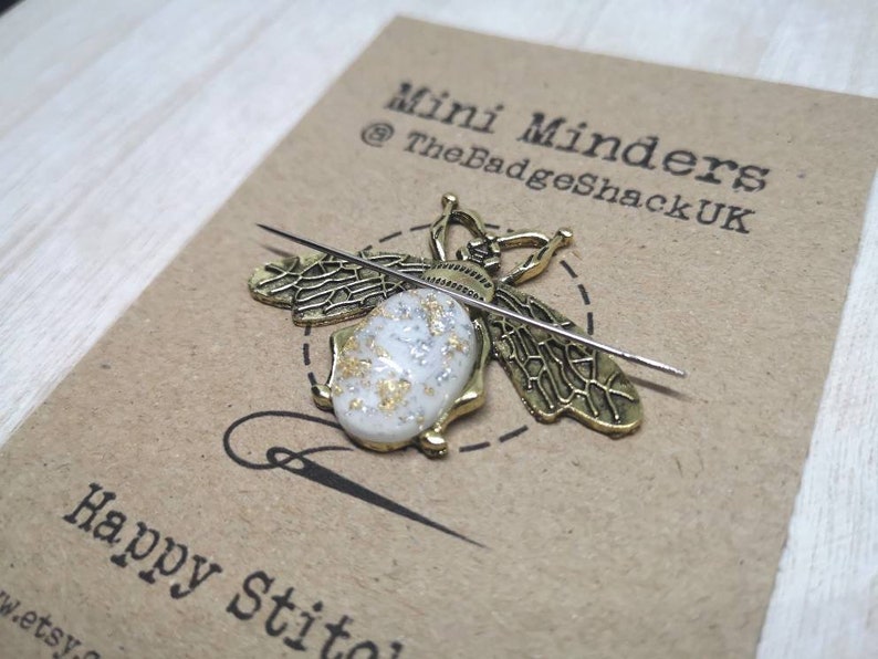 May include: A gold bee-shaped needle minder with a white and gold resin body. The needle minder is on a brown card with the text "Mini Minders @TheBadgeShackUK Happy Stitches".