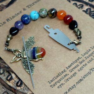 Op de afbeelding: Een naaldenmagneet en draadhouder met een messing bijenontwerp en een regenboogkleurige steen. De magneet is verbonden met een kralenketting van edelstenen, waaronder amethist, lapis lazuli en tijgeroog. De tekst "Needle Minder & Thread" is zichtbaar.