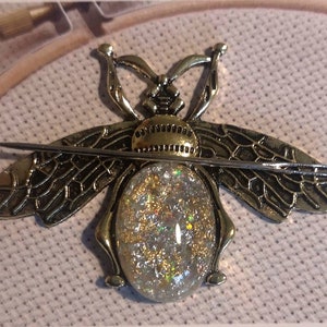 May include: A gold bee-shaped needle threader with a clear, sparkly, iridescent center. The threader is on a beige fabric background.