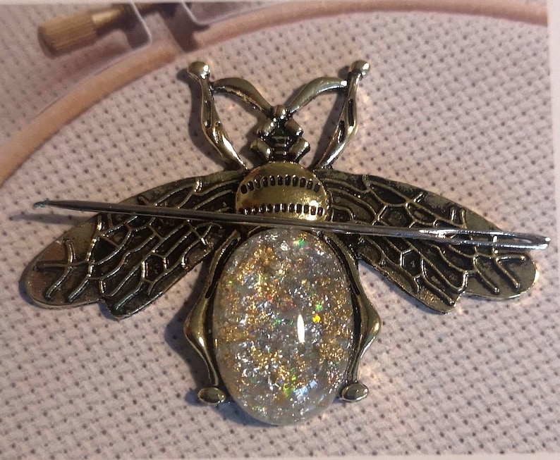 May include: A gold bee-shaped needle threader with a clear, sparkly, iridescent center. The threader is on a beige fabric background.