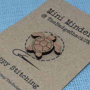 May include: A wooden turtle mini minder, a small wooden figure of a turtle, with the text "Mini Minders @TheBadgeShackUK" and "Happy stitching" on a brown card.