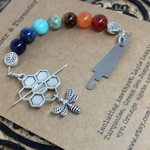 May include: A needle minder and threader set with a beaded bracelet. The bracelet has amethyst, lapis lazuli, turquoise, green sea jasper, tiger's eye, orange agate, and red agate beads. The set includes a silver-toned honeycomb and bee design.
