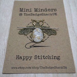 May include: A gold bee-shaped needle minder with a white and gold speckled center. The needle minder is on a brown card with the text "Mini Minders @TheBadgeShackUK" and "Happy Stitching www.etsy.com/shop/TheBadgeShackUK".