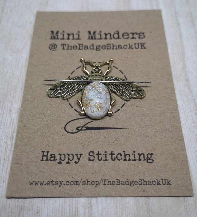 May include: A gold bee-shaped needle minder with a white and gold speckled center. The needle minder is on a brown card with the text "Mini Minders @TheBadgeShackUK" and "Happy Stitching www.etsy.com/shop/TheBadgeShackUK".