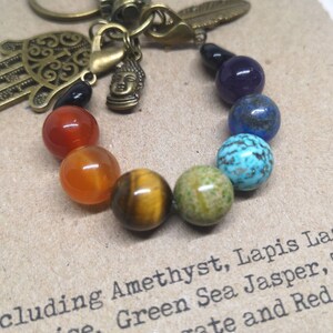 May include: A rainbow chakra bracelet with a hamsa hand charm, a Buddha charm, and a feather charm. The bracelet includes amethyst, lapis lazuli, green sea jasper, tiger's eye, and red agate beads.