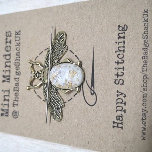 May include: Gold bee-shaped needle minder with a white and gold speckled resin body. The needle minder is attached to a thin metal needle. The card reads "Mini Minders @TheBadgeShackUK" and "Happy Stitching" with the website address "www.etsy.com/shop/TheBadgeShackUK".