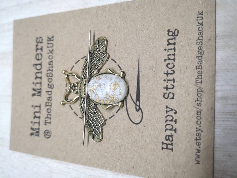 May include: Gold bee-shaped needle minder with a white and gold speckled resin body. The needle minder is attached to a thin metal needle. The card reads "Mini Minders @TheBadgeShackUK" and "Happy Stitching" with the website address "www.etsy.com/shop/TheBadgeShackUK".