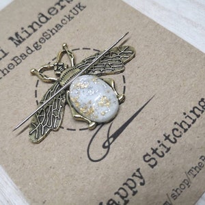 May include: A gold bee-shaped needle minder with a white and gold speckled body. The needle minder is on a brown card with the text "Mini Minder @TheBadgeShackUK" and "Happy stitching" with the shop name and website address.