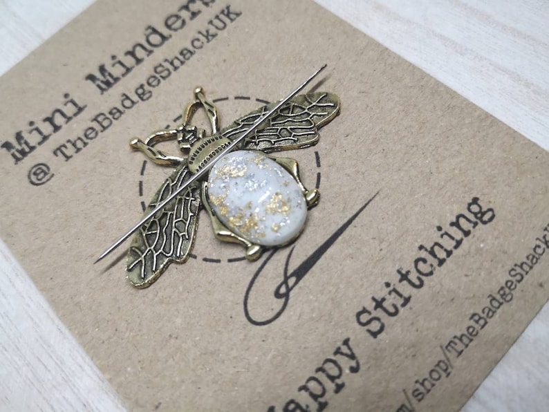May include: A gold bee-shaped needle minder with a white and gold speckled body. The needle minder is on a brown card with the text "Mini Minder @TheBadgeShackUK" and "Happy stitching" with the shop name and website address.