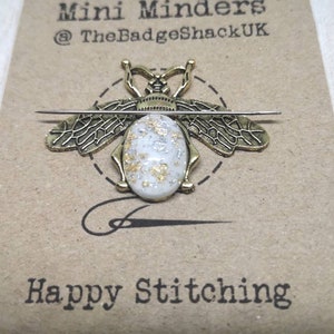 May include: Gold bee-shaped needle minder with a white oval center with gold and silver flecks. The needle minder is on a brown card with the text "Mini Minders @TheBadgeShackUK" and "Happy Stitching".