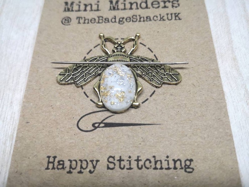 May include: Gold bee-shaped needle minder with a white oval center with gold and silver flecks. The needle minder is on a brown card with the text "Mini Minders @TheBadgeShackUK" and "Happy Stitching".