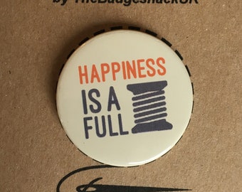 Może przedstawiać: Biały guzik z napisem "Happiness is a full" i szarą szpulką do szycia z pomarańczową nicią. Guzik znajduje się na brązowym tle z napisem "Needle Minder by TheBadgeshackUK" nad nim.