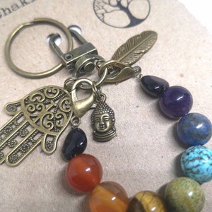 May include: A seven chakra keychain with a hamsa hand charm, a small Buddha charm, and a feather charm. The keychain is made with a variety of gemstones, including black onyx, amethyst, lapis lazuli, turquoise, tiger's eye, carnelian, and red jasper.