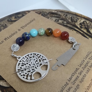 May include: A needle minder and threader with a silver tree of life design. The needle minder has a beaded strand of colorful gemstones. The text "Needle Minder & Threader" is visible.