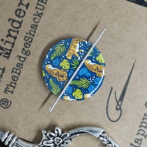 May include: A blue Mini Minder with a tiger and leaf design, with a silver needle. The text "Mini Minder @TheBadgeShackUK" is visible. Silver scissors are at the bottom. The design is circular.