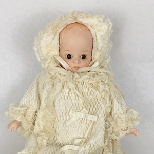 May include: A vintage baby doll dressed in a cream-colored lace gown and bonnet. The doll has a painted face with dark eyes and a slightly open mouth. The gown features a lattice design and is adorned with small bows.