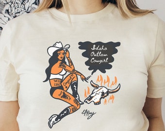 May include: A white t-shirt with a graphic of a woman in a cowboy hat, holding a cigarette and sitting on a skull with flames. The text "Idaho Outlaw Cowgirl" is in a speech bubble above her head. The graphic is signed "El Ray".