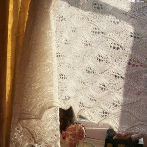 May include: A white crocheted shawl with a delicate lace pattern hangs over a window. The shawl is draped over a yellow curtain and a floral patterned fabric. A glass candle holder sits on a table in front of the window.