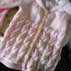 Peut inclure: Un cardigan rose en tricot avec un devant boutonné et un motif délicat en dentelle. Le cardigan a six boutons blancs.