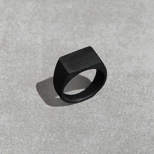 Könnte beinhalten: Ein schwarzer Siegelring mit rechteckiger Oberseite. Der Ring hat eine gebürstete Oberfläche.