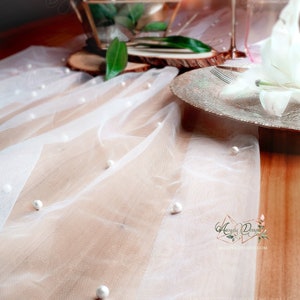 May include: White tulle table runner with pearl accents. The runner is on a wooden table with a glass terrarium, a white flower, and a silver plate.
