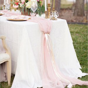 May include: A white tablecloth with a pink chiffon table runner and gold candlesticks. The table is set with a floral centerpiece and a white chair.