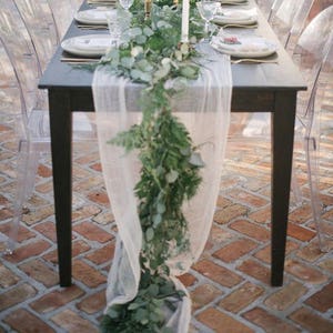 May include: A long, rectangular table with a white tablecloth and a garland of greenery down the center. The table is set with white plates, gold silverware, and clear glasses. There are candles in gold holders down the center of the table. The table is surrounded by clear ghost chairs.
