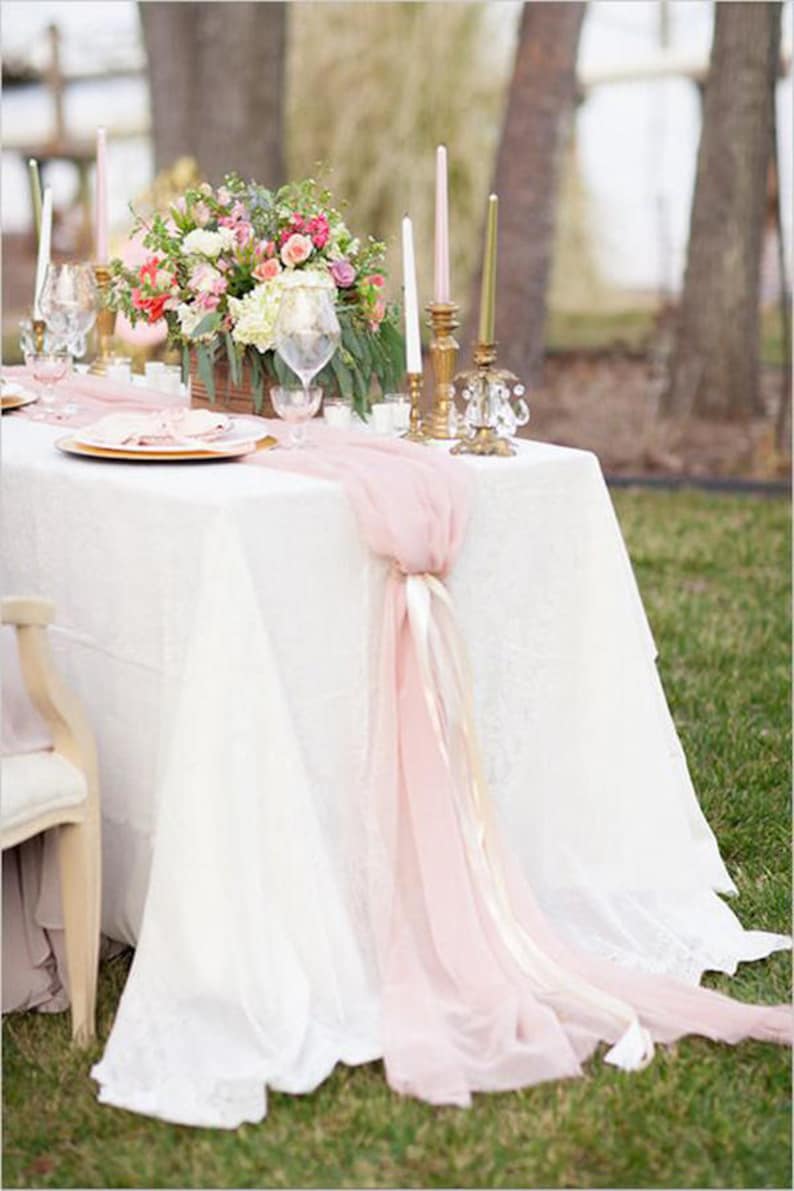 May include: A white tablecloth with a pink chiffon table runner and gold candlesticks. The table is set with a floral centerpiece and a white chair.