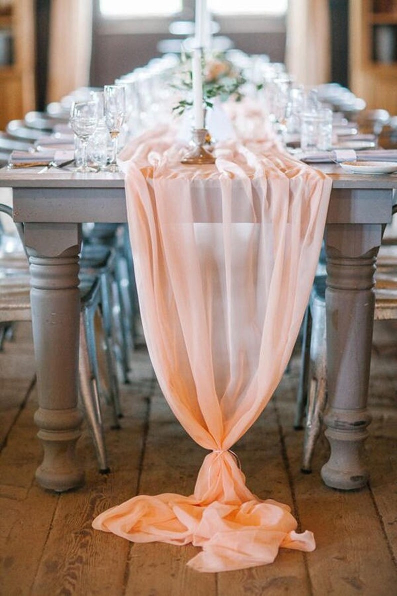 May include: A long, peach-colored sheer fabric table runner drapes over a rustic wooden table with white paint. The runner is gathered at the center and hangs down to the floor. The table is set with white plates, silverware, and glasses.