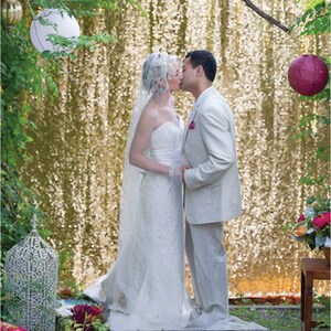 May include: A bride and groom kiss in front of a gold sequin backdrop. The bride is wearing a white strapless wedding dress with a veil. The groom is wearing a light gray suit. There are pink and white paper lanterns hanging from a tree branch above the couple.