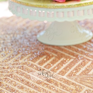 May include: A two-tiered cake stand with a gold rim and a white base. The stand is decorated with a delicate scalloped edge. The stand is sitting on a rose gold sequin tablecloth.