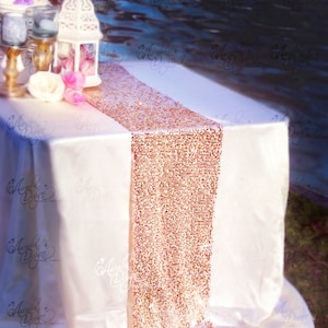 Op de afbeelding: Een witte tafel met een roze gouden sequin tafelkleed. De tafel is gedekt met een wit tafelkleed en een lantaarn.