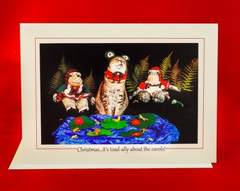 Könnte beinhalten: Weihnachtskarte mit einem getigerten Kater und zwei Froschfiguren, alle in festlicher Kleidung. Das Bild enthält Seerosenblätter und den Text "Christmas...it's toad-ally about the carols!"