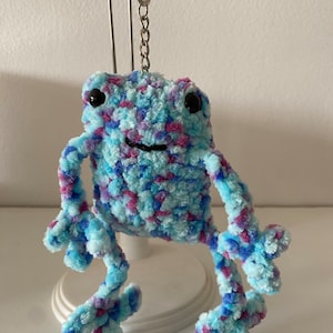May include: A blue, purple, and pink yarn frog keychain with black eyes. The frog is sitting on a white stand.