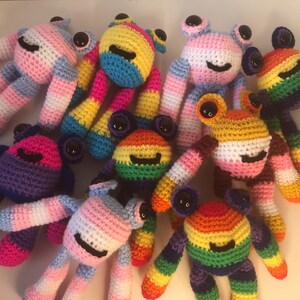 Pode incluir: Uma coleção de peluches de sapo de crochê em várias cores, incluindo arco-íris, rosa, azul e amarelo. Cada sapo tem olhos grandes e pretos e uma cara sorridente.