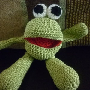 May include: Green crocheted frog toy with white eyes and a red mouth. The frog is sitting on a brown and green striped fabric.