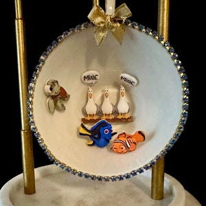 May include: A white porcelain teacup with a blue rhinestone trim, featuring a scene of three seagulls with the word "MINE" above each, a blue tang fish, an orange clownfish, and a green sea turtle. The teacup is on a gold stand with a white marble base.