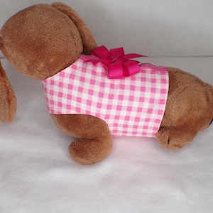 May include: A brown plush bunny wearing a pink and white gingham patterned shirt with a pink bow. The bunny has long ears and is lying down on a white surface. The shirt is a sleeveless design.
