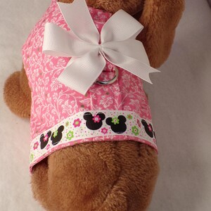 May include: Pink dog harness with a white floral pattern and a white bow. The harness has a decorative band with black mouse silhouettes and floral accents. A silver metal ring is attached for leash attachment.